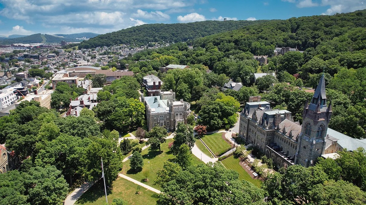 Aerial view of a university campus with historic buildings, lush greenery, and a prominent church with a tall steeple, surrounded by a city and rolling hills in the background.