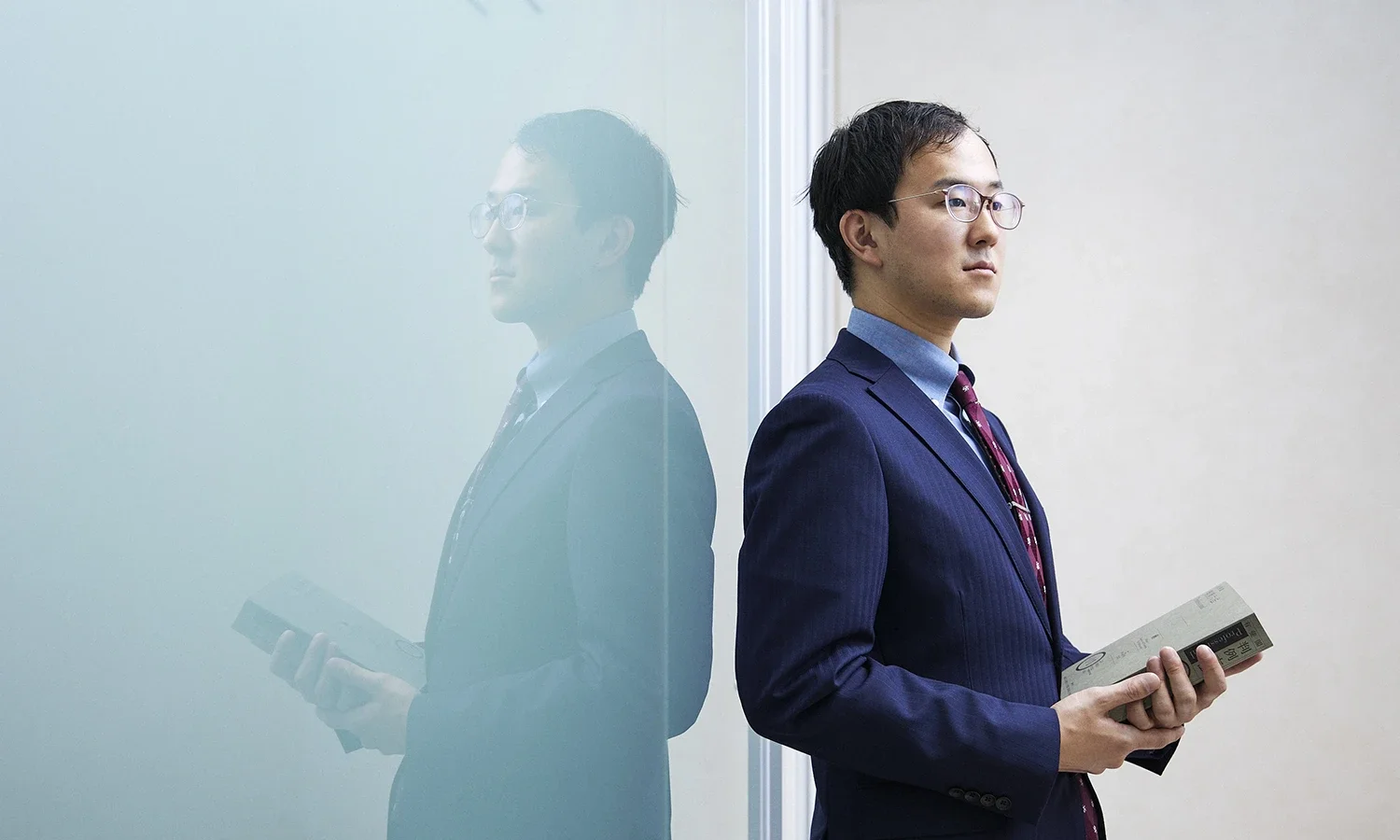 スーツを着た男性がコップを持ちながら壁にもたれて立っている