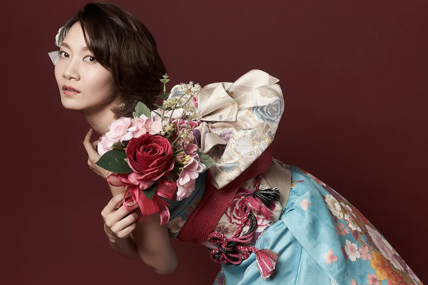 和服を着た女性が花束を持ち、横向きにポーズをとっている。背景は深赤色。
