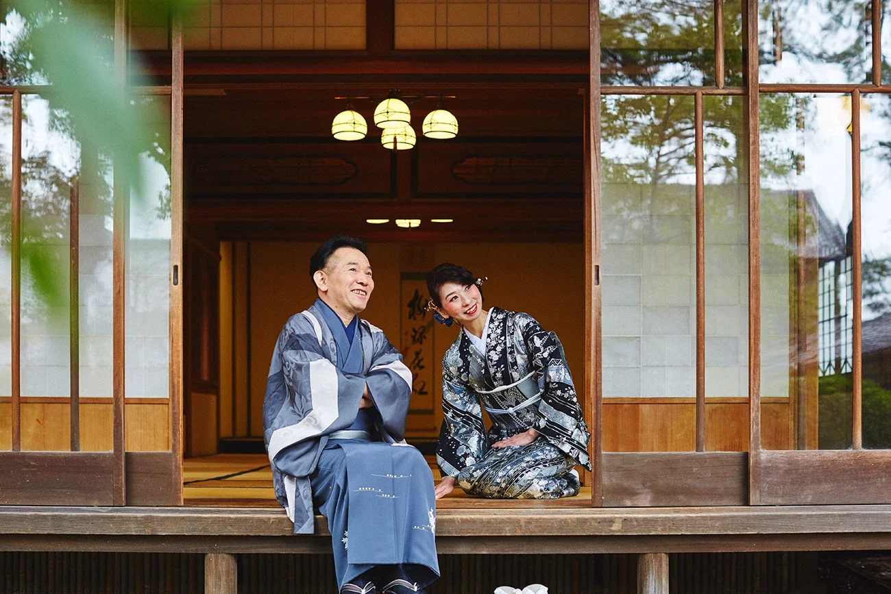 伝統的な日本の建物の縁側で、着物を着た男性と女性が座っている。男性は青色の着物を着ており、女性は黒と銀の花柄の着物を着ている。