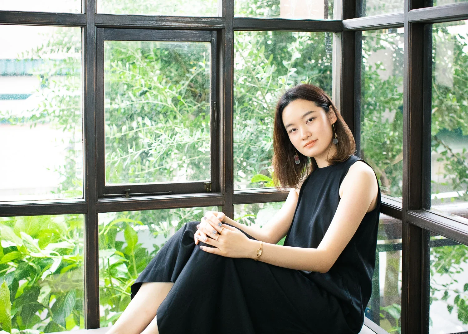 黒い服を着た女性が窓際に座り、緑の植物が見える背景の前でポーズをとっている。