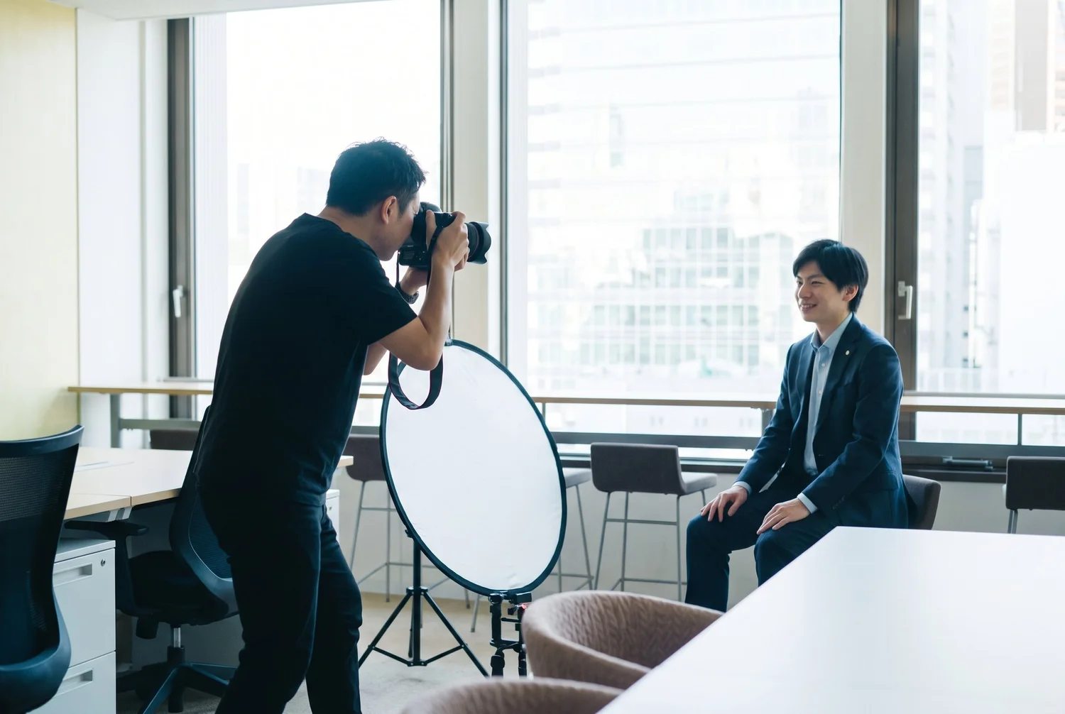 オフィスの会議室で、窓からの自然光とレフ板を使って社員のビジネスポートレートを撮影するカメラマン