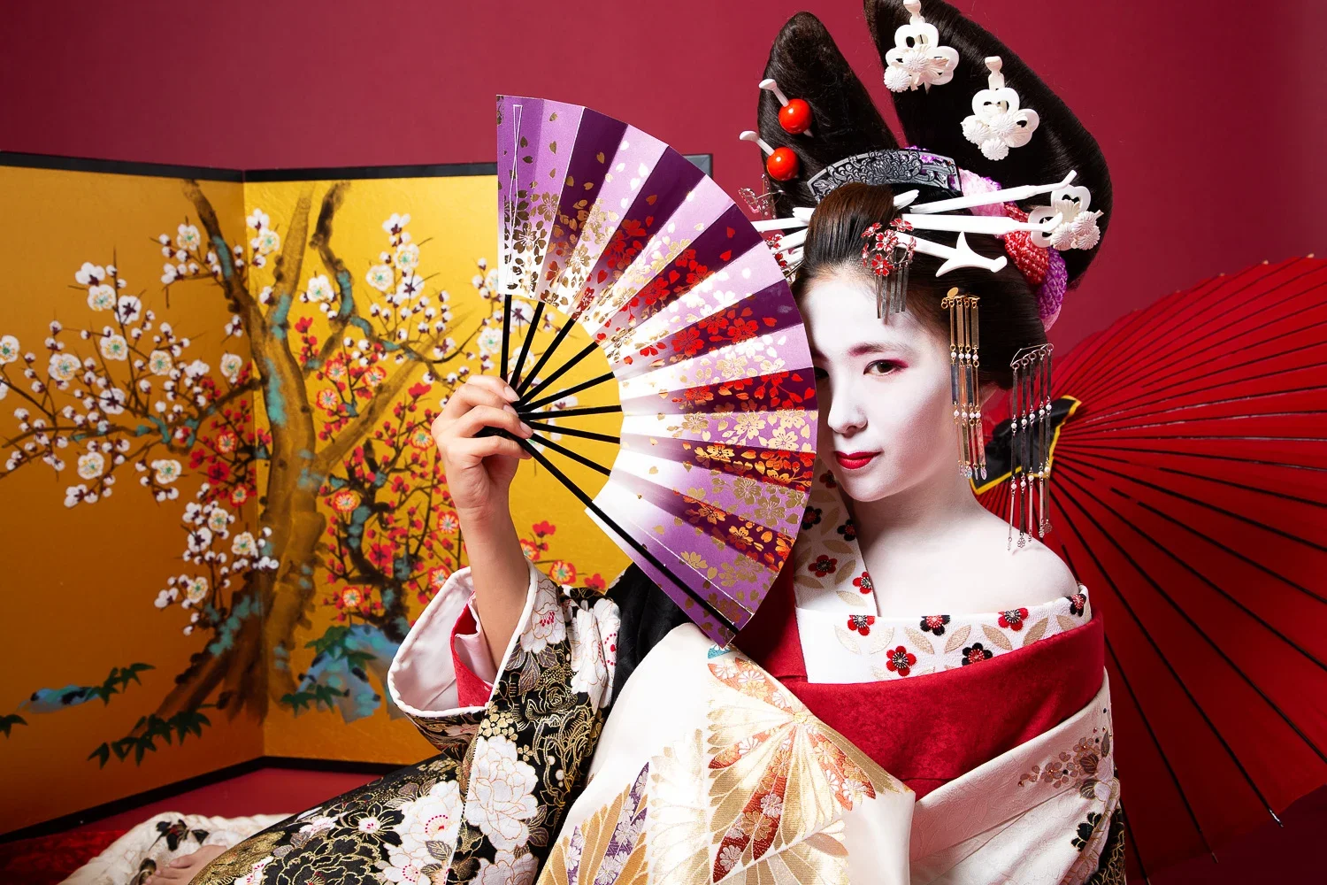 着物を着た女性が花柄の折りたたみ扇子を持ち、着物と髪飾りに和のデザインを身にまとい、背景に金色の桜の絵が描かれた屏風が見える。