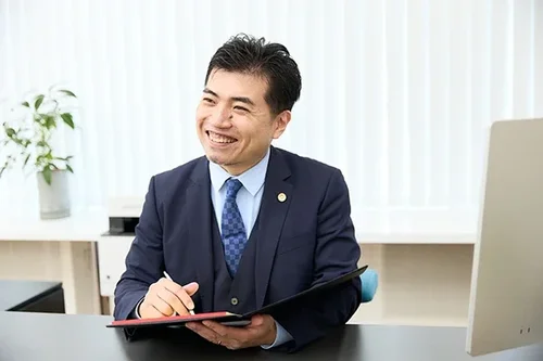 ベンナビなどのポータルサイトに最適な、親しみやすい笑顔の弁護士宣材写真。事務所のデスクで手帳を手に相談に乗る日本人弁護士。