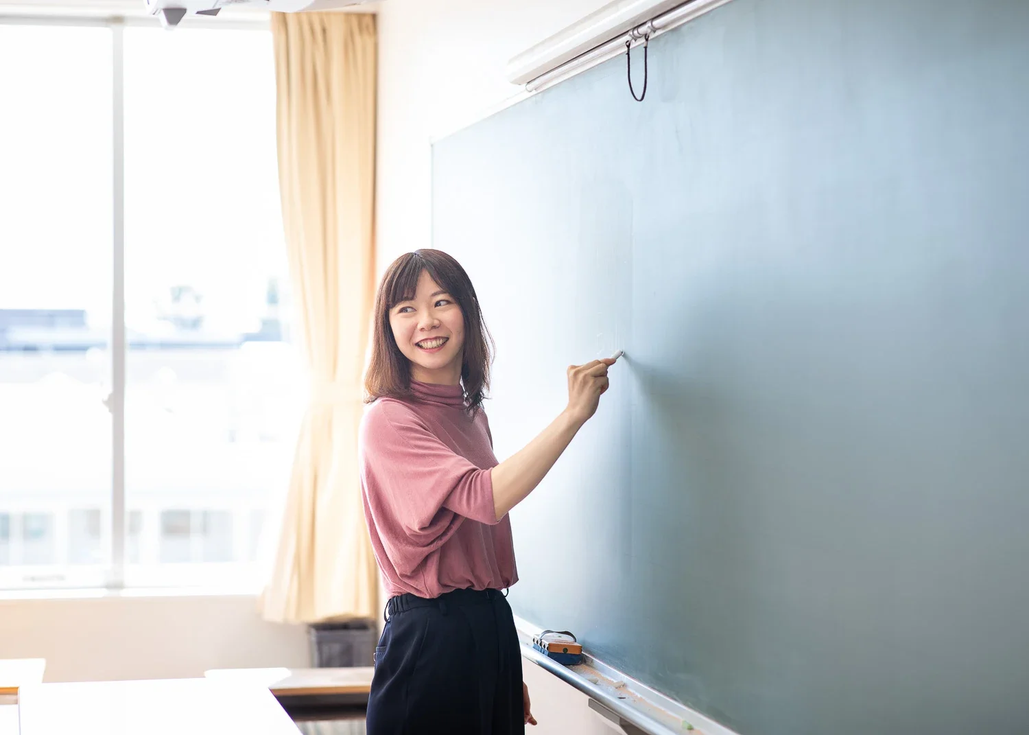 黒板の前で笑顔の女性教師がチョークを持って立っている教室の内観