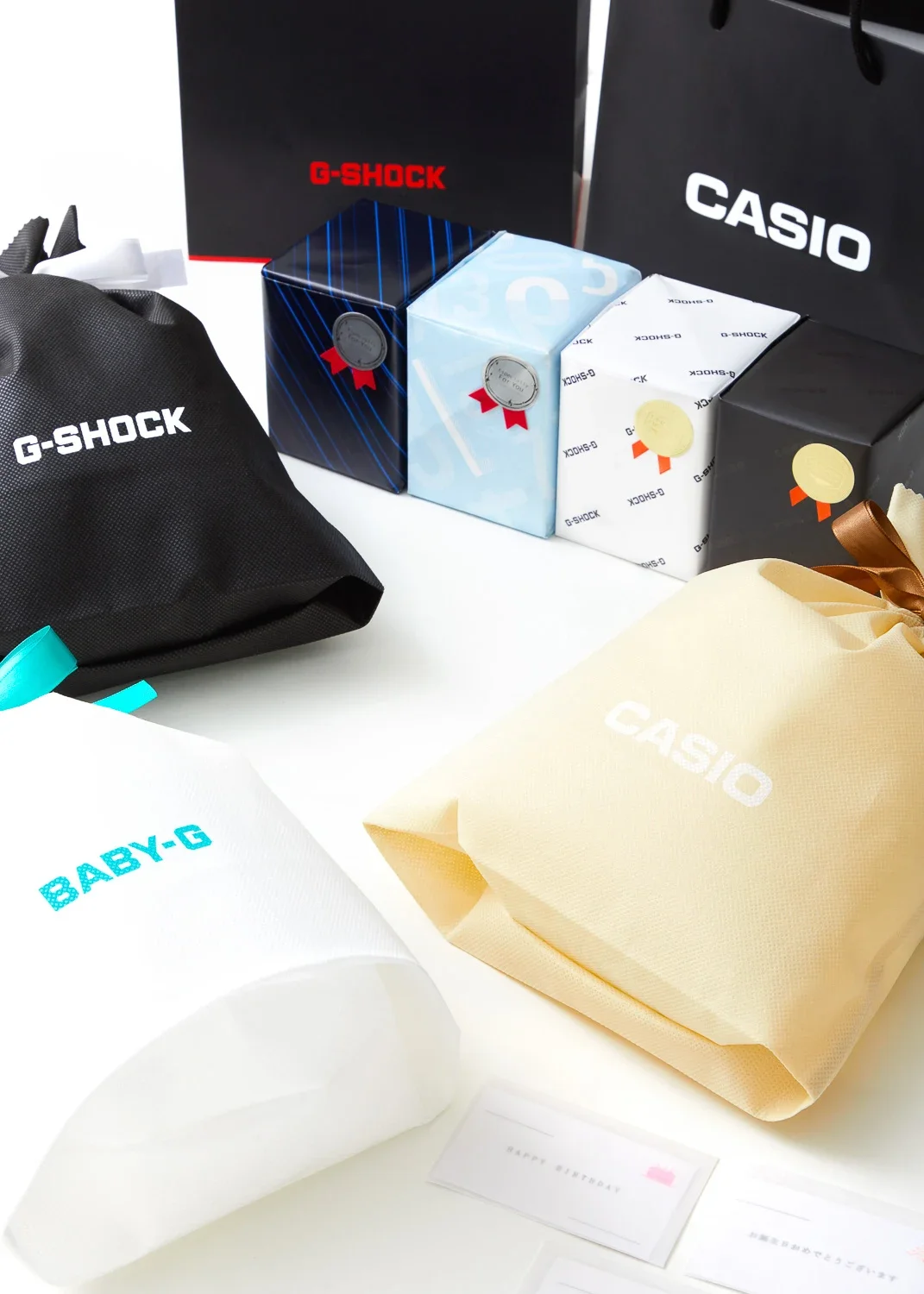 ギフトバッグとボックス、黒や白、青、黒の箱にG-SHOCKやCASIOのロゴが見える時計のパッケージ、バースデーカードも見える。