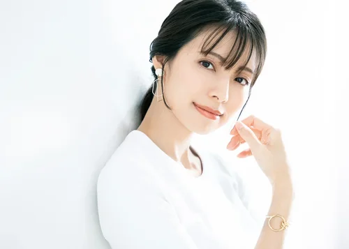 白い背景の前で微笑む若い女性。長い黒髪で耳にイヤリングをつけている