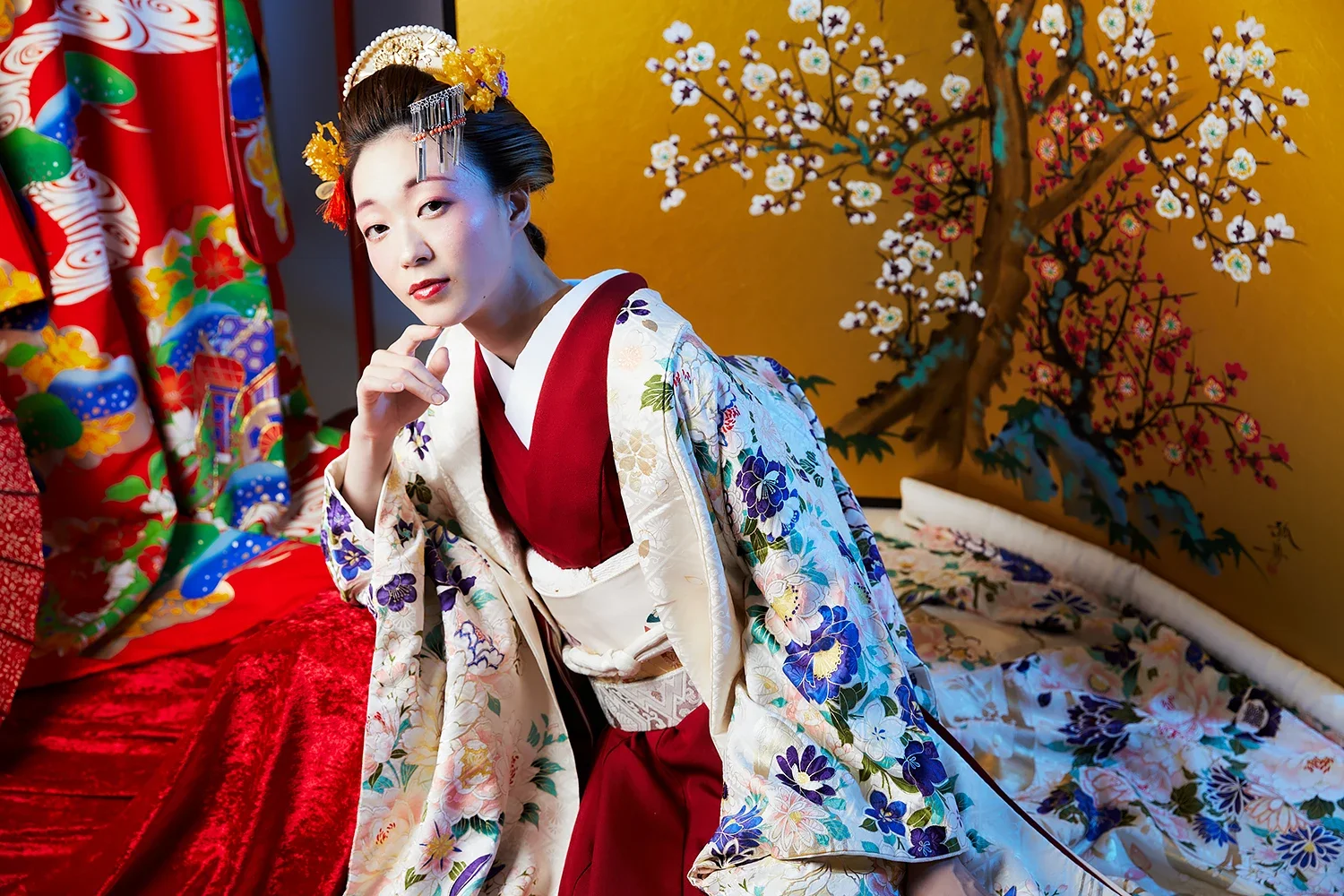 伝統的な着物を着た女性が髪飾りをつけてポーズをとっている。背景には金色の屏風と、桜の絵が描かれた屏風が見える。