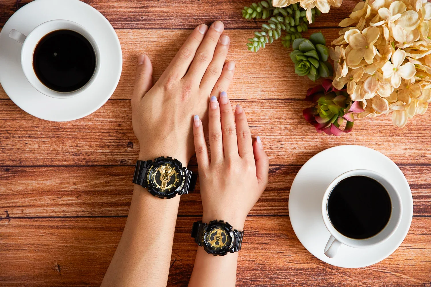 木製のテーブルの上に、2つの白いコーヒーカップとソーサー、2つの腕と手、腕時計、そして花束が置かれている写真です。