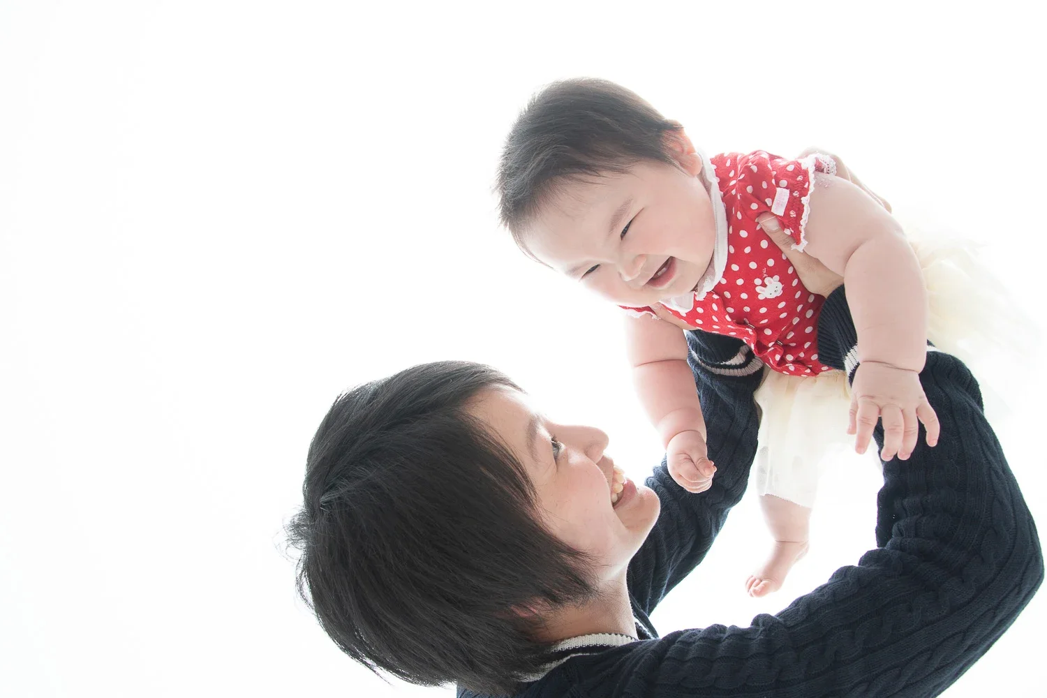 笑顔の女性が赤いドット柄のシャツを着た赤ちゃんを持ち上げている写真