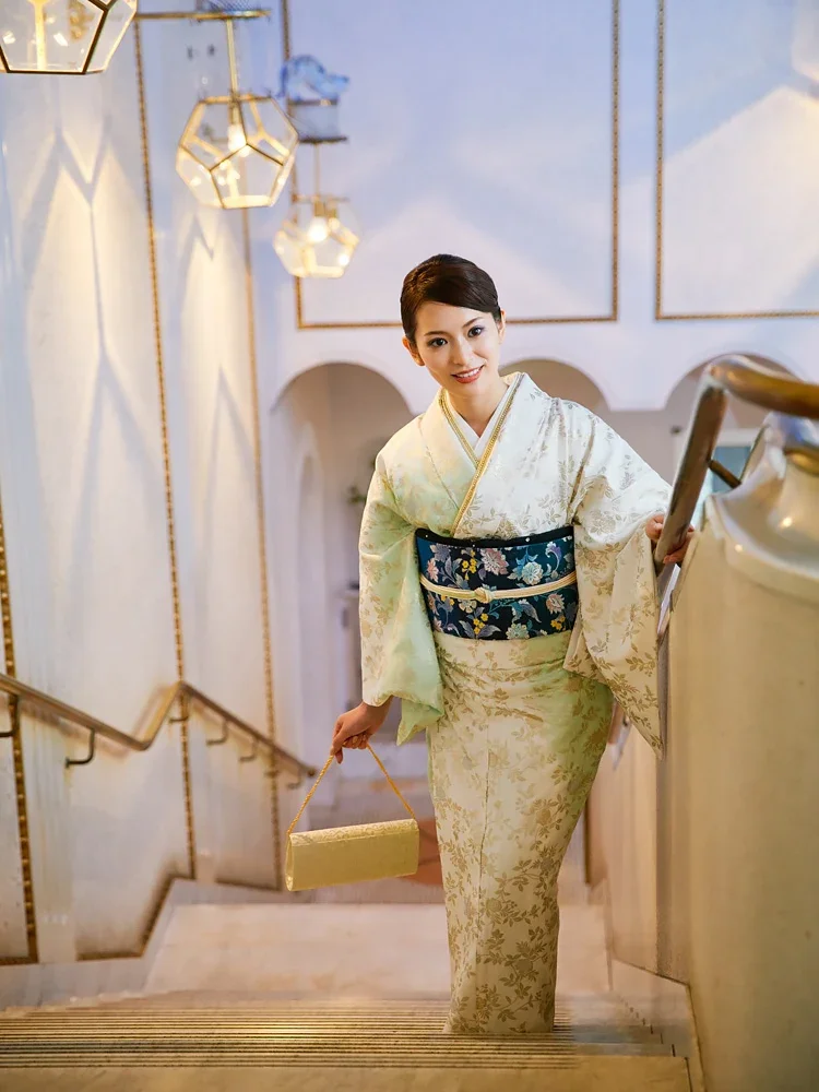和服を着た若い女性が階段を上がっている。彼女は笑顔でクラッチバッグを持っている。豪華な照明と装飾がある室内空間。