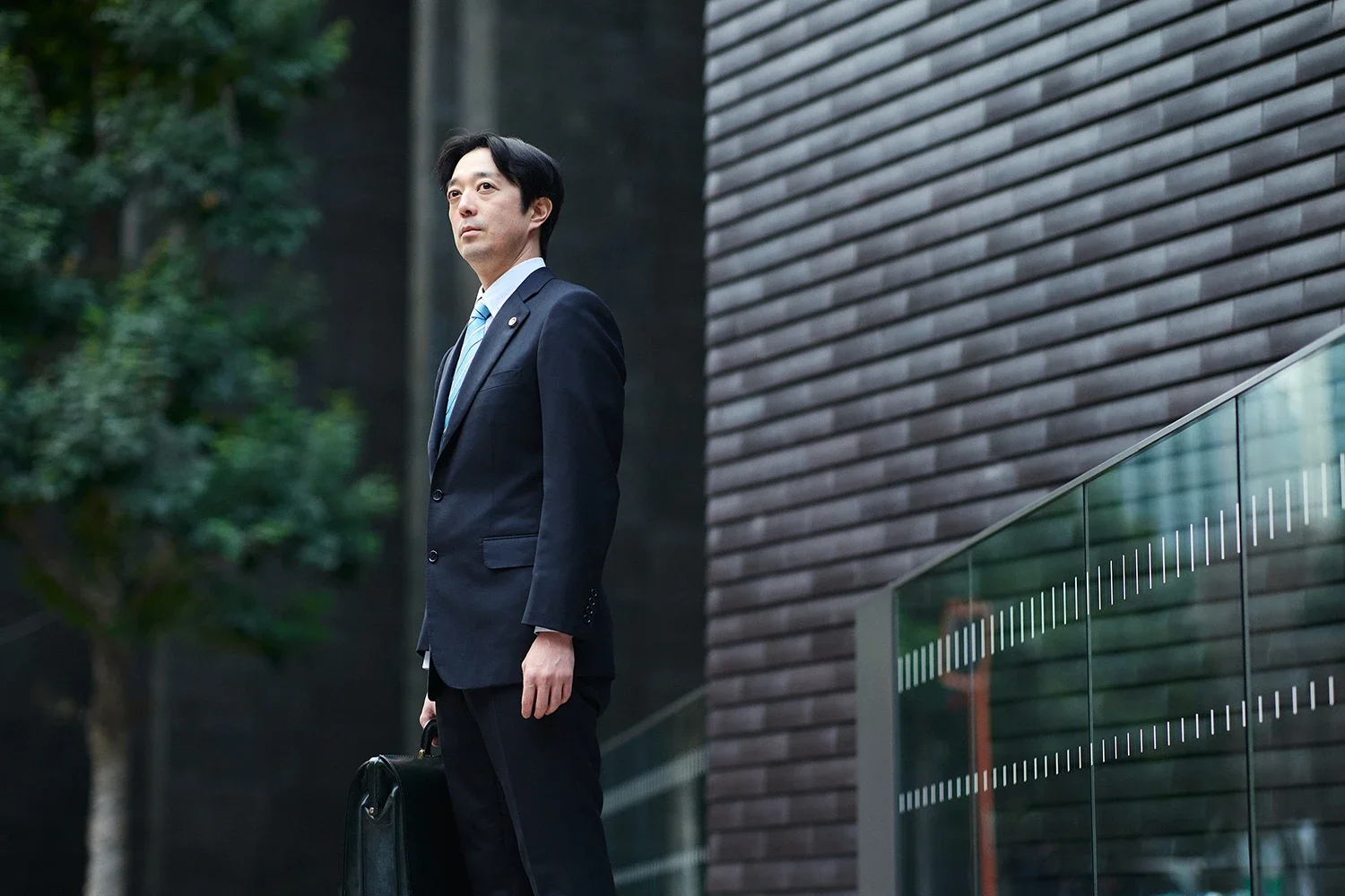 ビジネスマンがスーツを着て、トートバッグを持ちながら建物の外に立っている写真