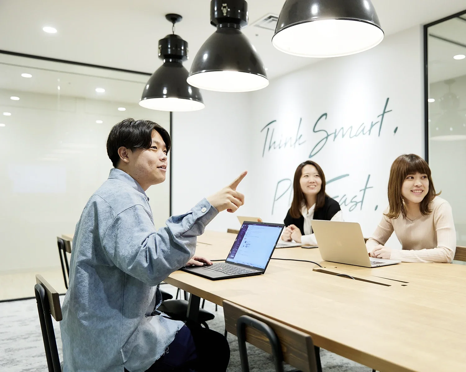 ビジネスミーティングをしている若い男女のグループ。白い壁に「Think Smart, Think Fast」と書かれている会議室。三人の女性と一人の男性が座っている。