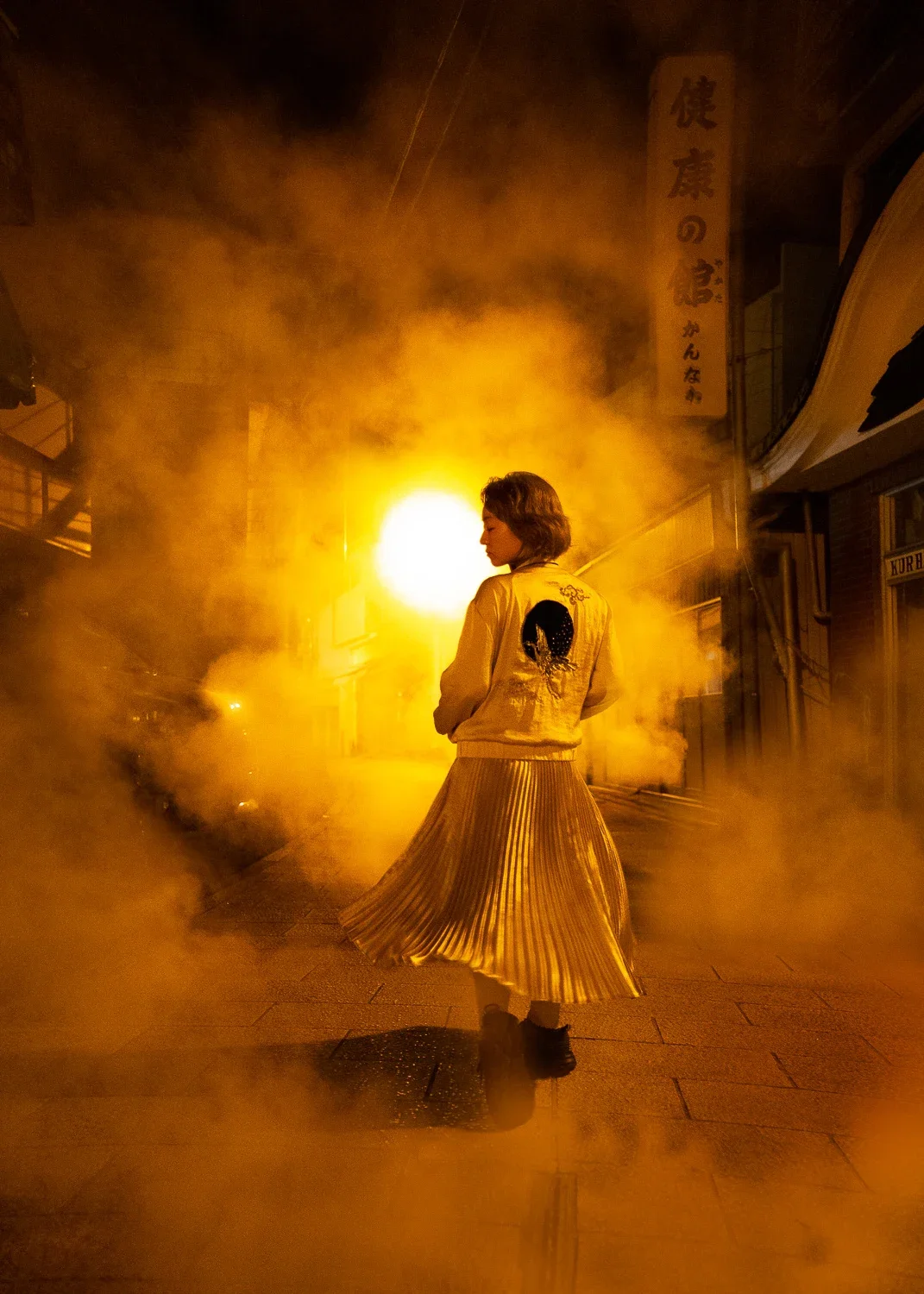 夜の街で女性が煙の中を歩いている。背景に明るい黄色い光と看板が見える。