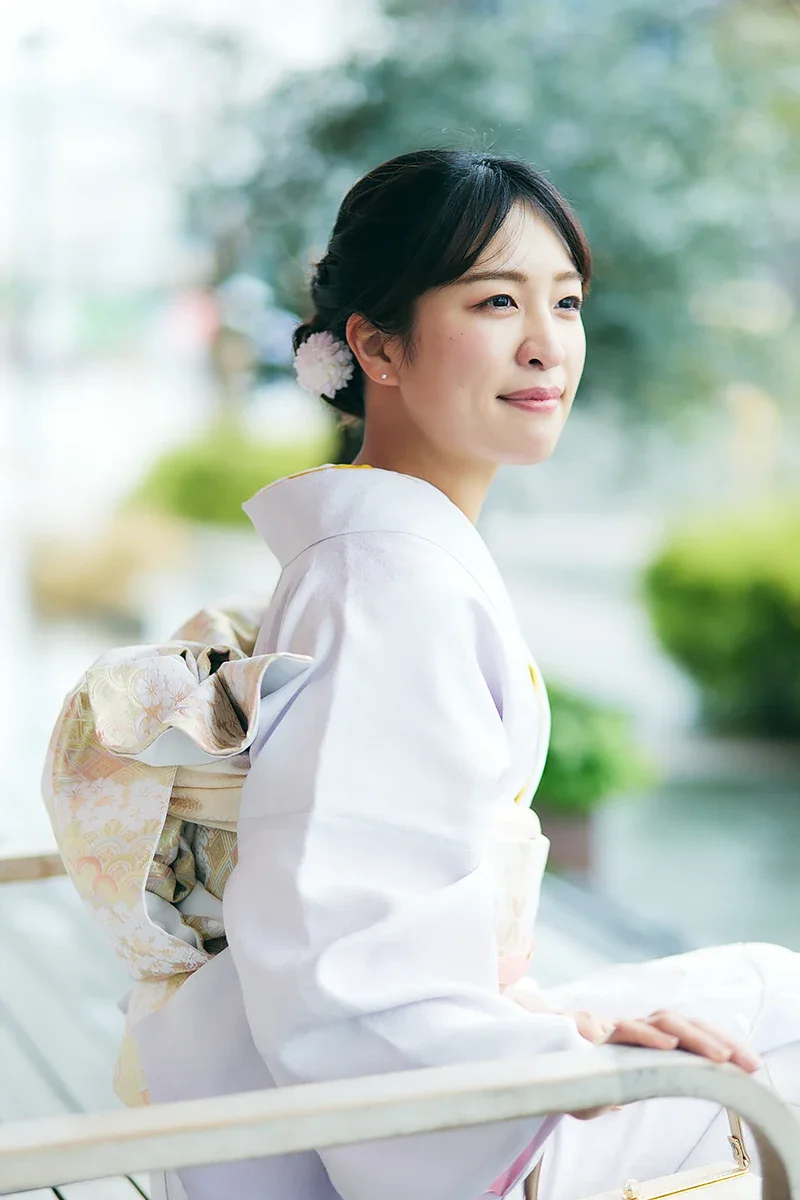 白い着物を着た若い女性が座っている写真。背景はぼかされている。女性は微笑みながら景色を見ている。」}