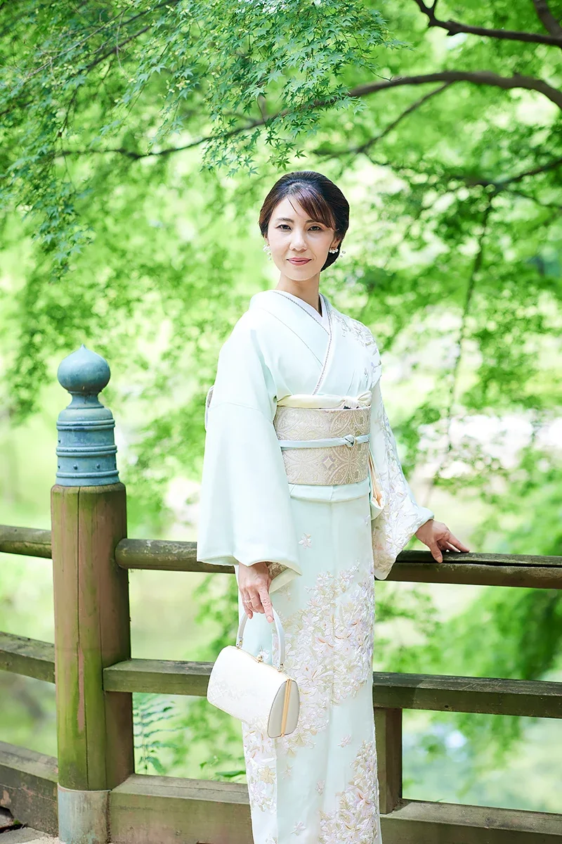 緑豊かな公園の橋の柱のそばで白い着物を着た女性が微笑んでいる写真