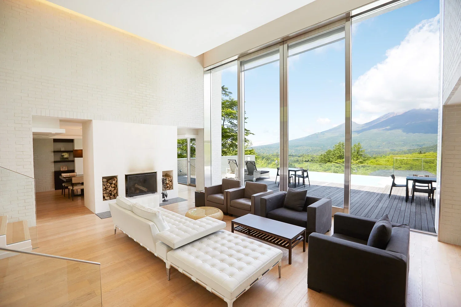 山と自然が見える大きな窓があるリビングルーム。ソファや椅子、暖炉が配置されているモダンなインテリア。木製のフローリングと白い壁。屋外のデッキにはテーブルと椅子がある。