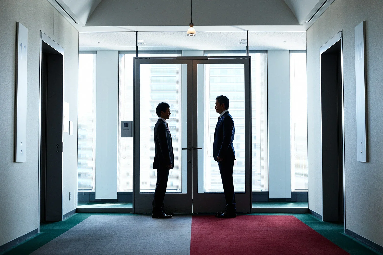 ビルのロビーで向き合って話す二人のビジネスマン。背後に大きな窓とエレベーターのドアが見える。