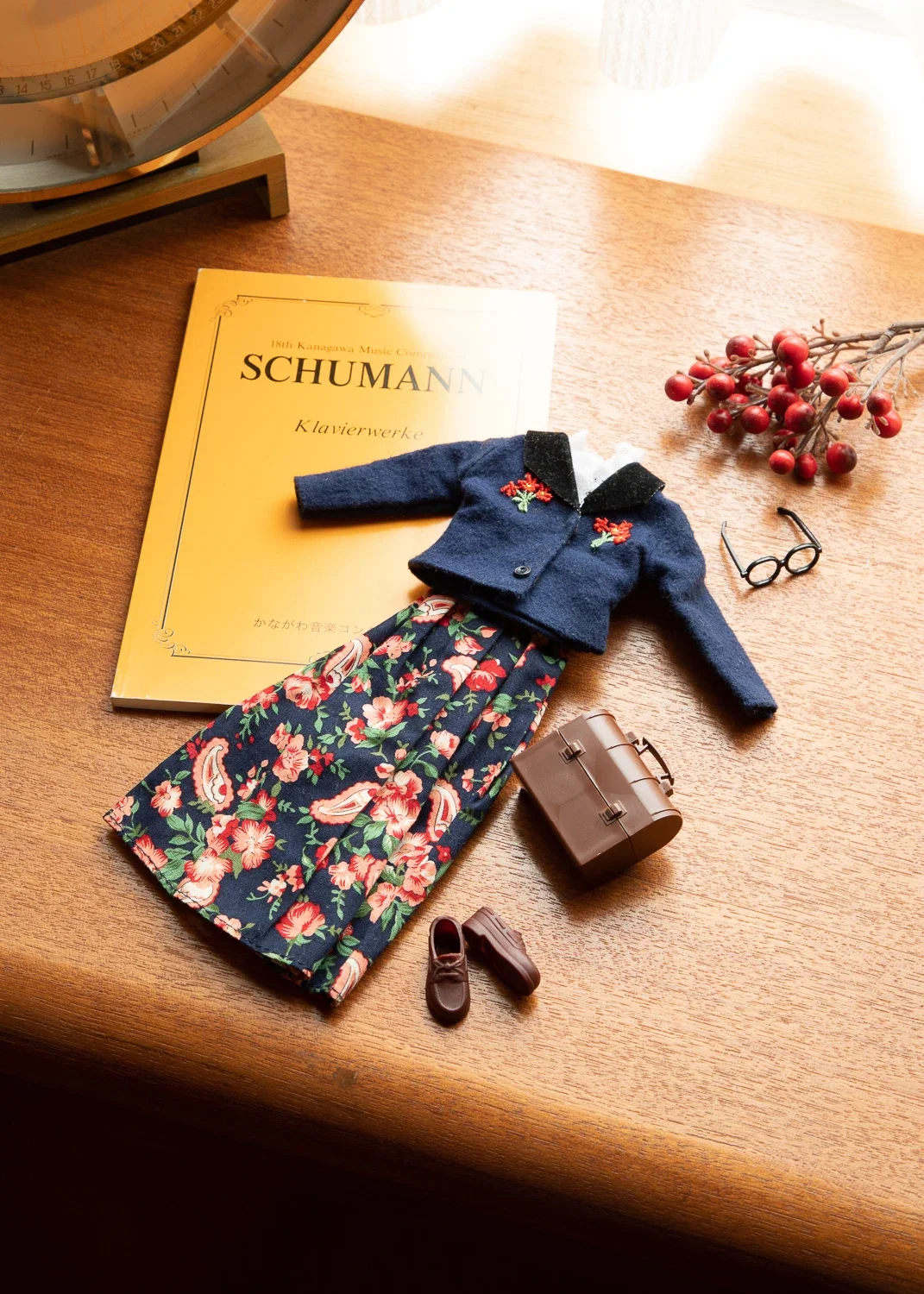 木の机の上に、楽譜の本、花柄のワンピース、セーター、眼鏡、ミニチュアのブーツと小さなケース、赤い実のついた枝が置かれている。