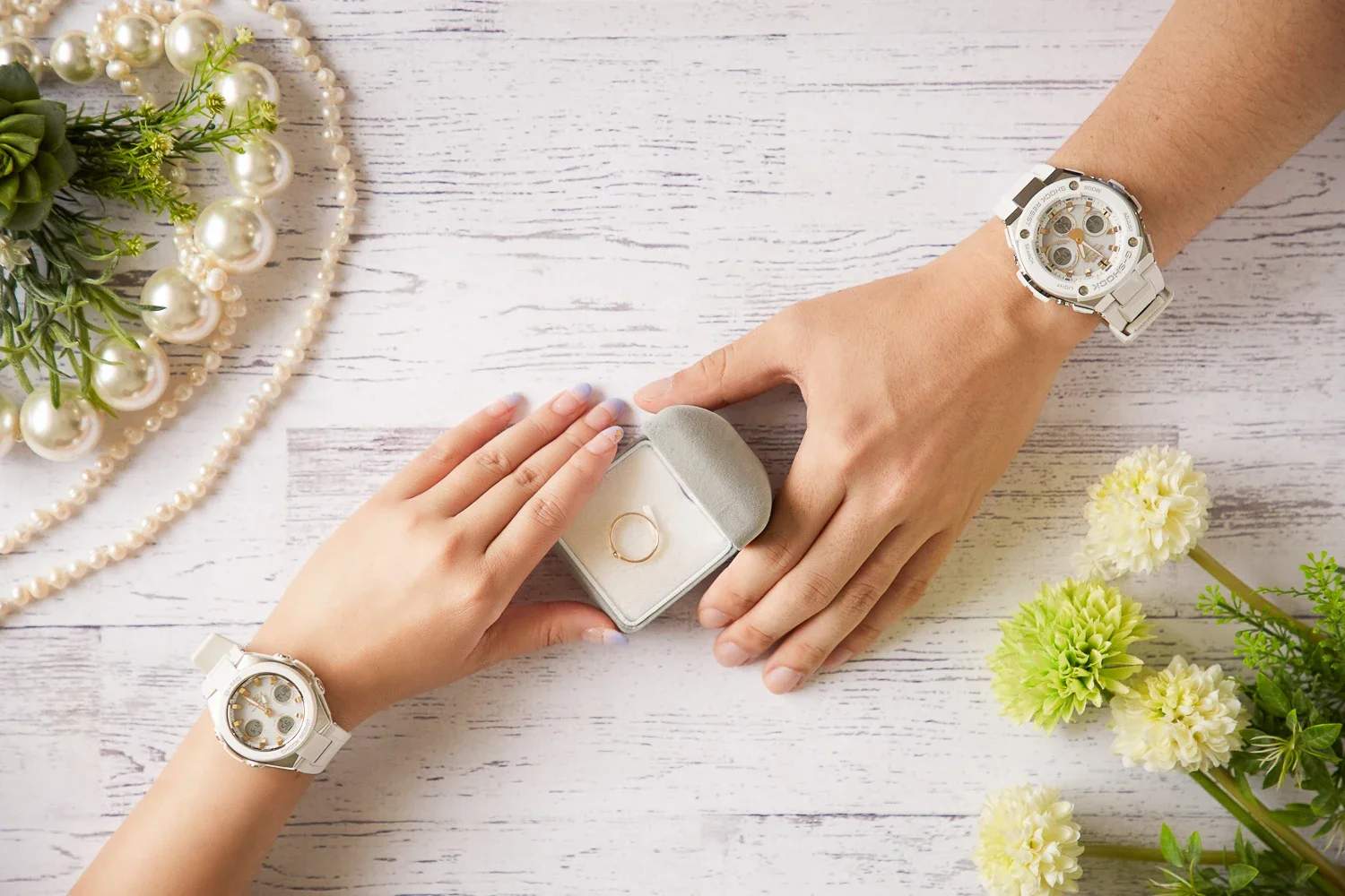両手に似た白い腕時計を着けた人がお互いに結婚指輪を渡している。周囲に花とパールのネックレスが置かれた白い木目調のテーブルの上の場面.