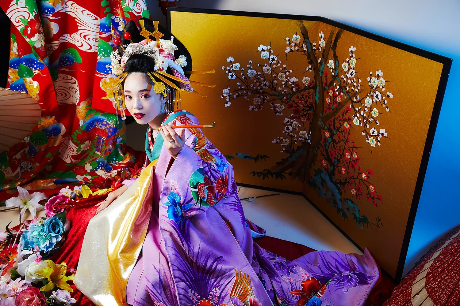 伝統的な着物を着た女性と、花や扇子、背景の屏風に梅の花の絵が描かれた装飾的な室内の写真