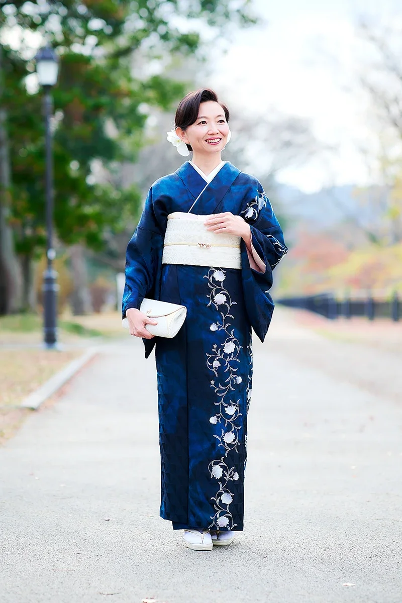 青い着物を着た女性が笑顔で歩いている写真です。背景には木と公園のような場所があります。