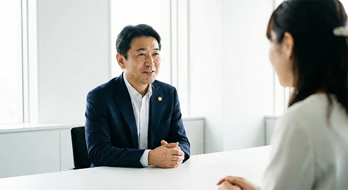 白基調の明るい会議室で、女性の相談者（手前・後ろ姿）の話を穏やかな笑顔で聞いている男性弁護士の撮影イメージ。自然光の中で親しみやすさと安心感を伝える相談イメージ。
