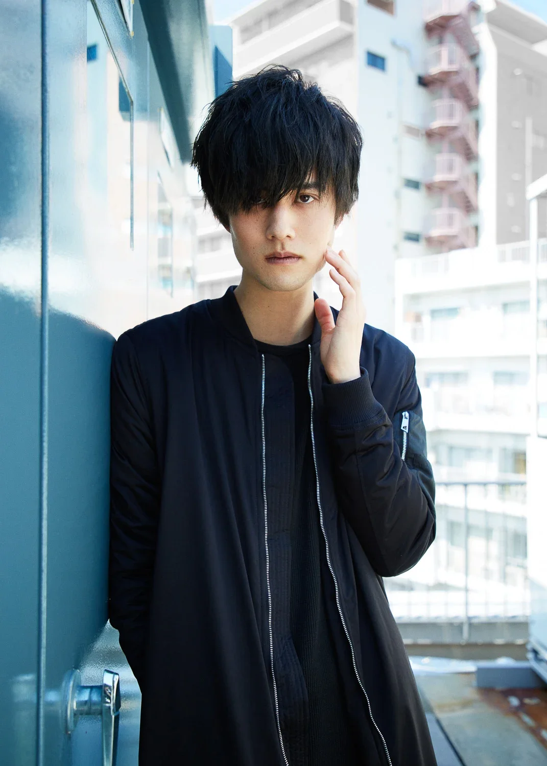 黒いジャケットを着た若い男性が壁に寄りかかって立っている。背景は都市の高層ビルの外観。