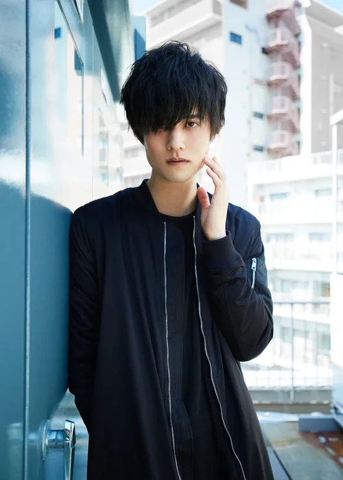 黒いジャケットを着た若い男性が壁に寄りかかって立っている。背景は都市の高層ビルの外観