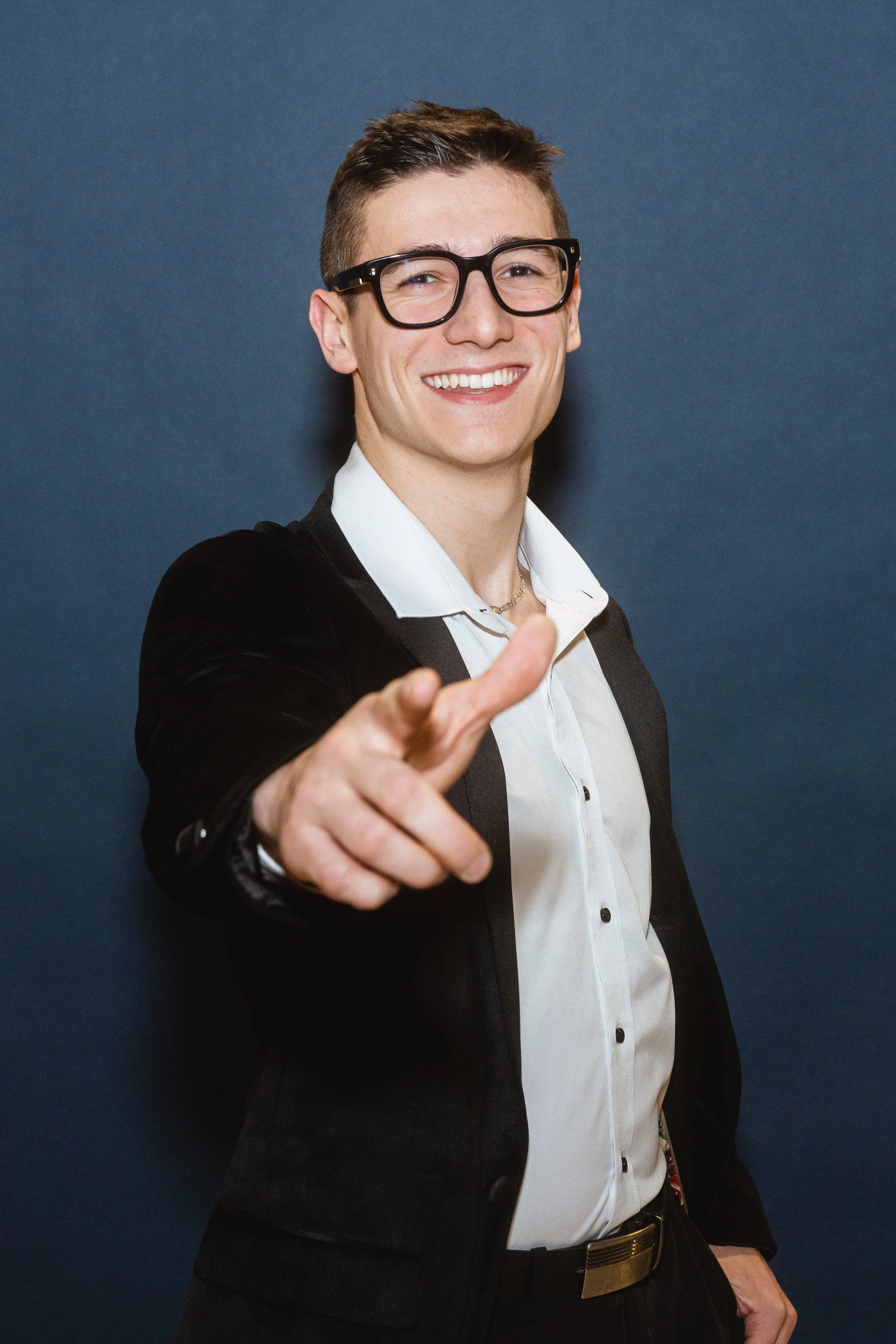 Jeune homme souriant portant des lunettes noires, vêtements formels, pointant du doigt vers l'objectif, debout contre un mur bleu.