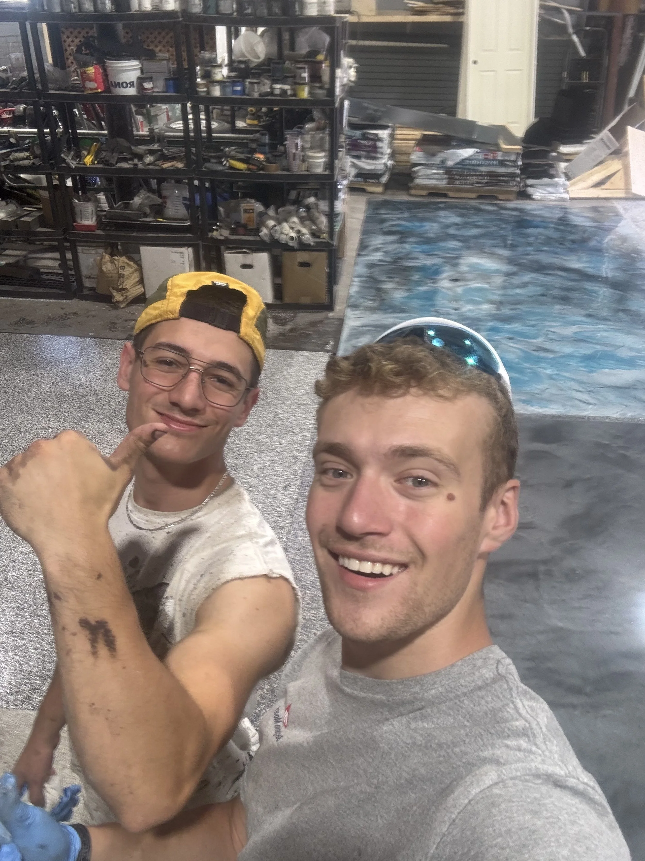 Deux jeunes hommes prennent une selfie dans un atelier, en arrière plan des planchers d'époxy métallique et de polyaspartic flakes