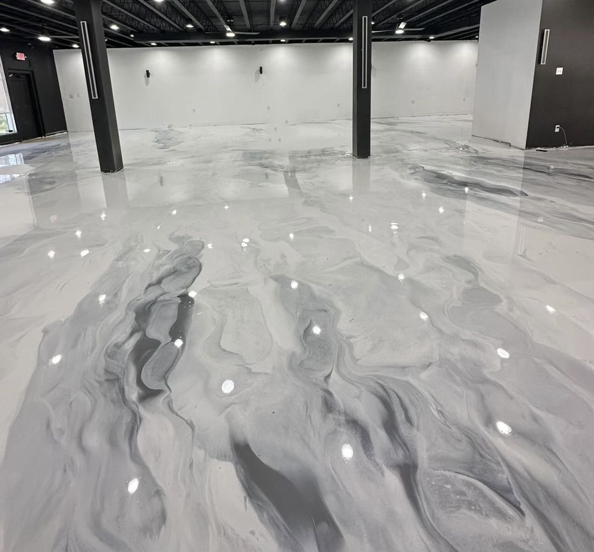 Salle vide avec un sol en époxy métallique blanc et gris à motifs, murs gris clair, colonnes noires, plafond noir avec éclairage intégré.
