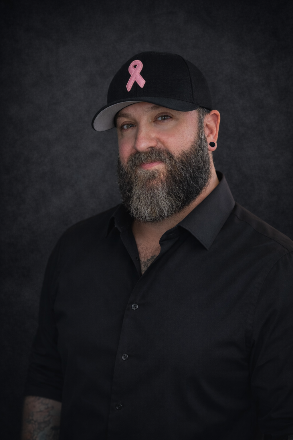 A portrait photo of Sean Brow, an areola reconstructive tattoo artist and coach, wearing a black baseball cap with a pink cancer ribbon symbol and a black button-up shirt, standing against a dark background.