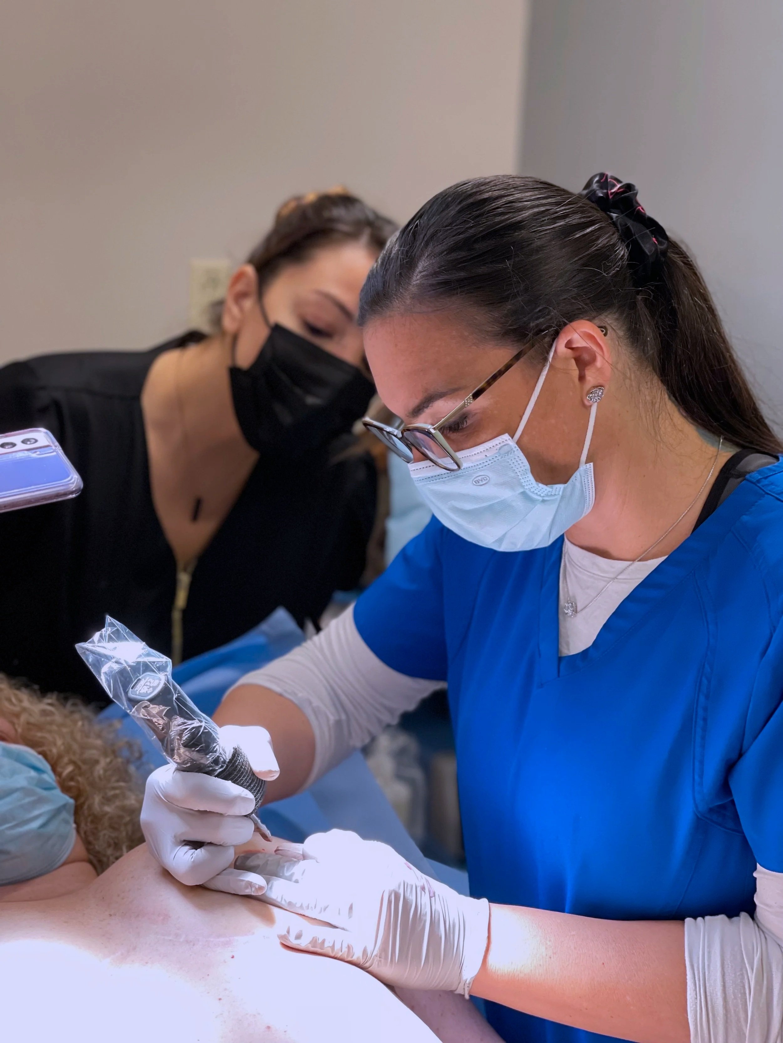 Kara Gutierrez, the areola reconstructive tattoo artist, dressed in blue scrubs and glasses performs a tattoo procedure on a patient while another person observes, both wearing masks.
