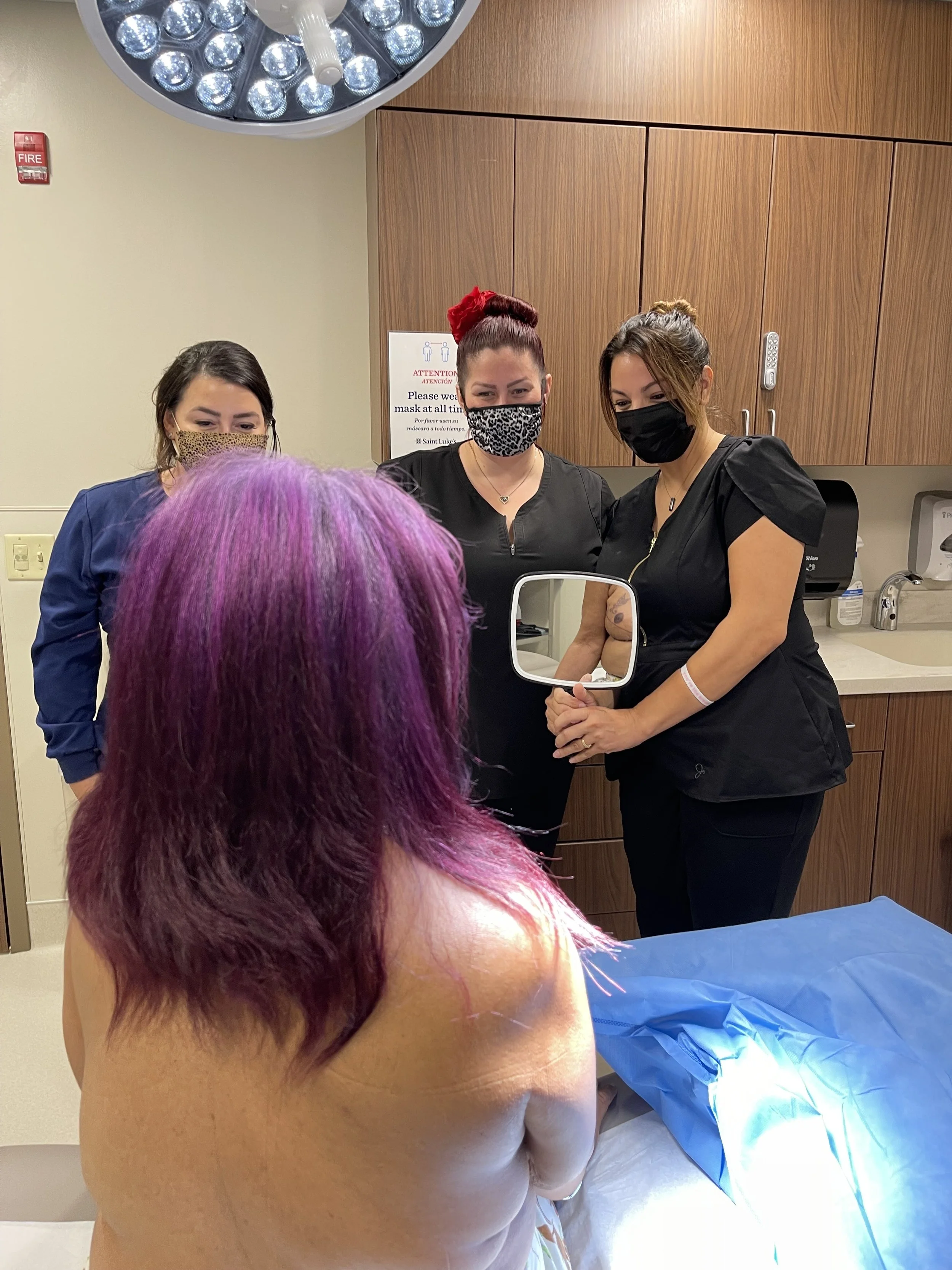 A patient with purple hair sitting on an examination table in a medical setting, with areola reconstructive tattoo students observing.