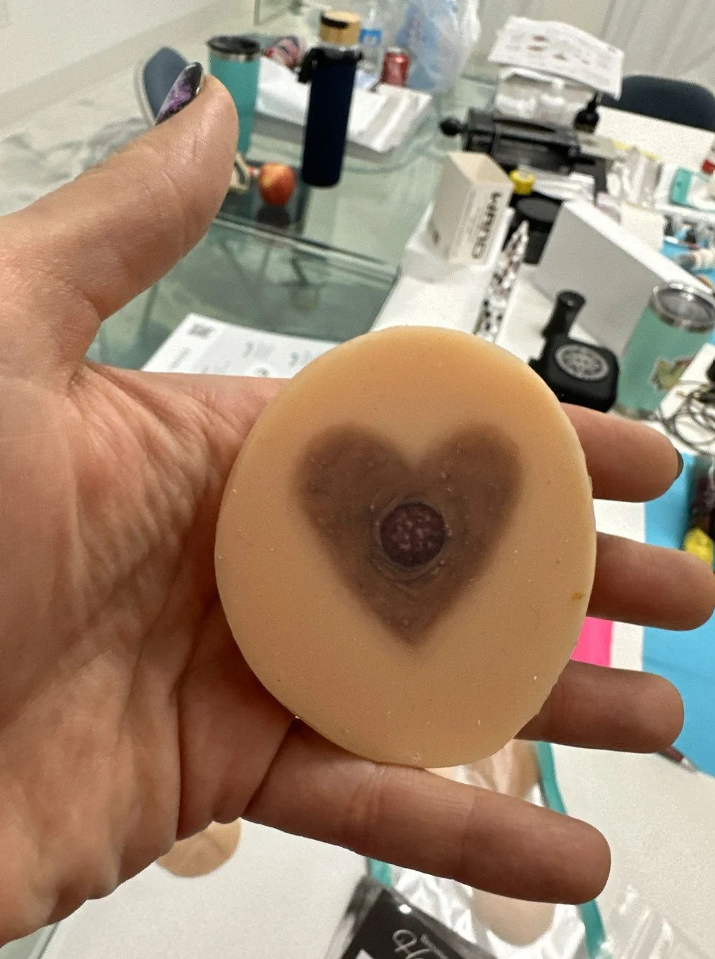 A hand holding a flesh-colored silicone object with a dark, heart-shaped areola design tattooed at the center. The background shows a class table with various items including bottles, papers, and tattoo supplies.