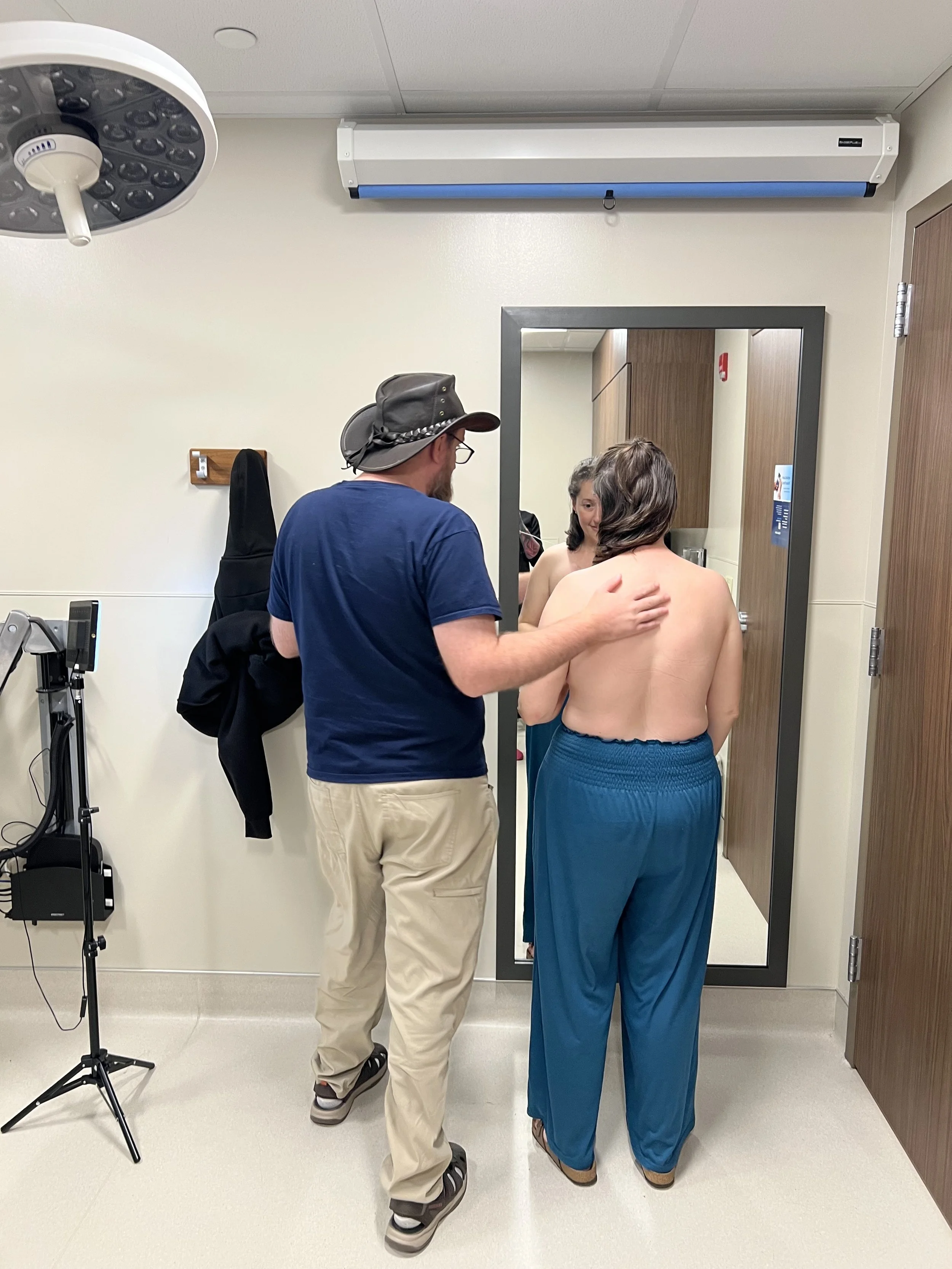 A husband and wife lovingly holding each other's arm while examining the final results of the wife's reconstructive areola tattoo in a medical office with a mirror, medical equipment, and a light fixture on the ceiling.