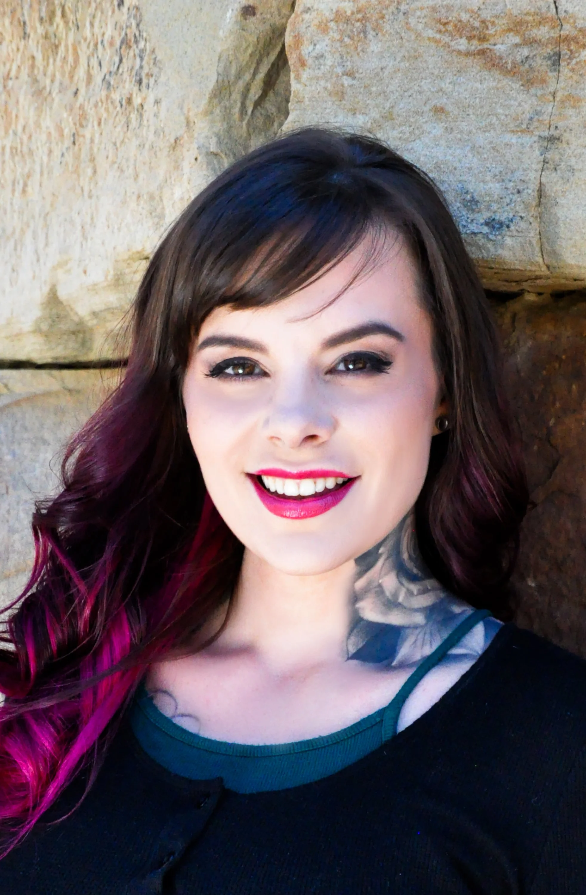Portrait photo of Liz Cook Tattoo, smiling with dark brown and pink hair with pink tips, wearing a black top with a visible teal strap, standing against a stone wall.