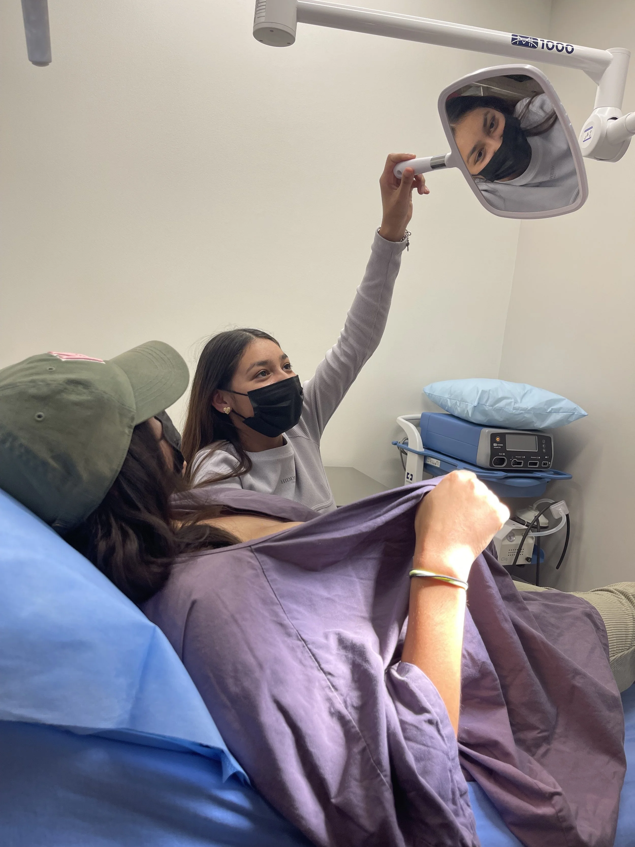A woman in a hospital bed wearing a black face mask, with a woman standing next to her wearing a gray shirt and black face mask, adjusting a mirror to see a reflection of the woman's freshly tattooed reconstructive areola.