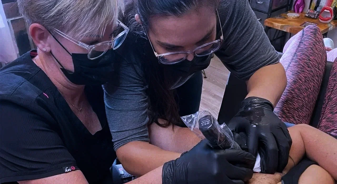 Kara Gutierrez, an areola reconstructive tattoo instructor and student wearing masks and glasses tattooing a reconstructive areola tattoo in a class setting.