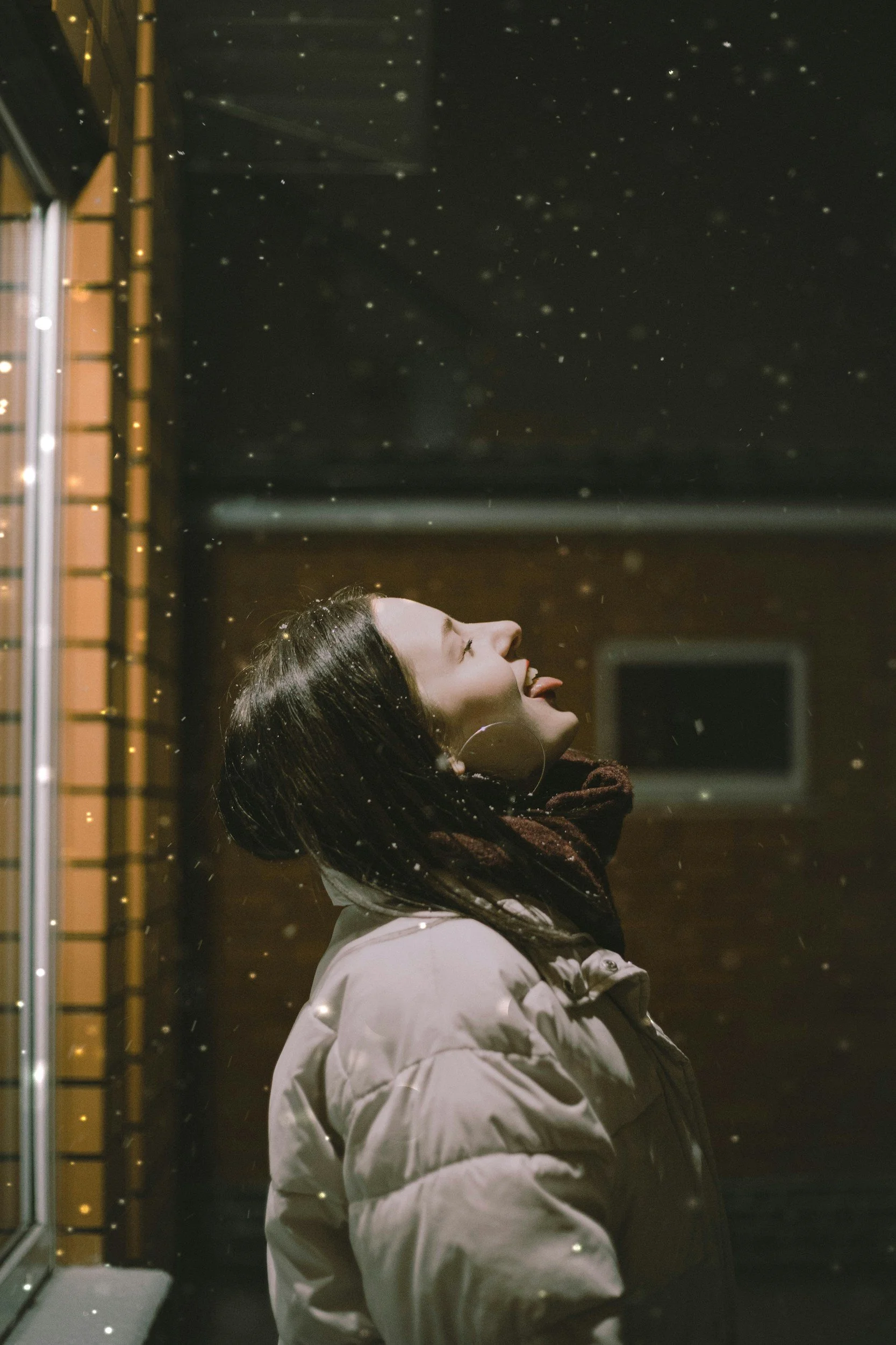 A woman in a beige jacket standing outdoors at night with snow falling around her, smiling and sticking her tongue out.