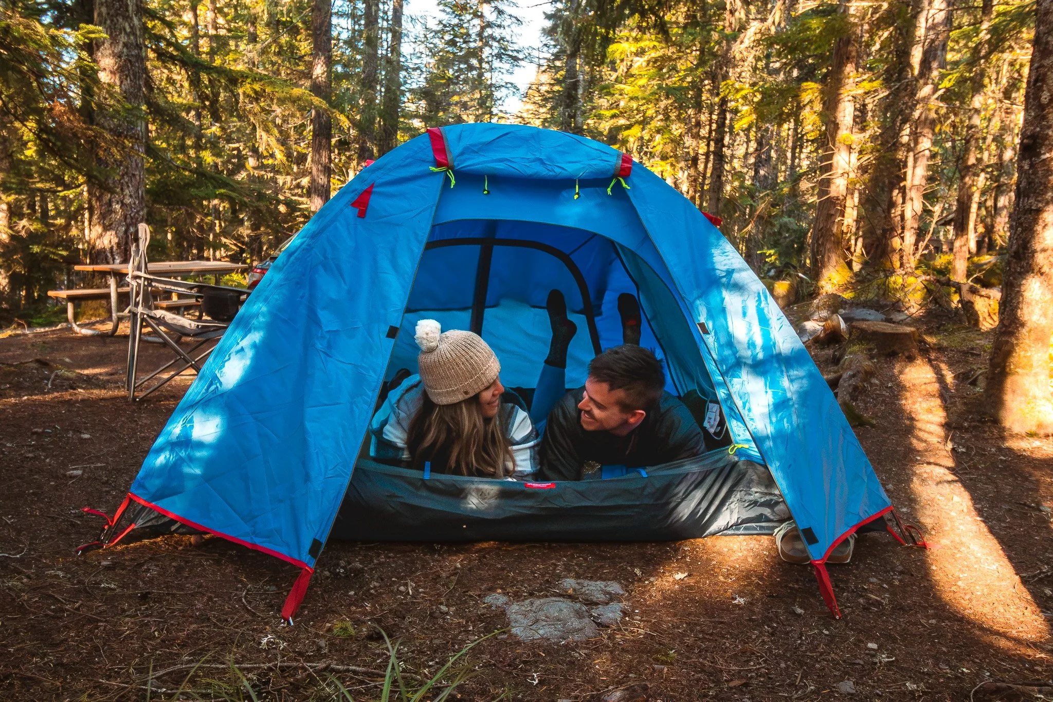 Couple lying inside a blue tent smiling at each other, feet propped up, campsite in a sunlit forest.