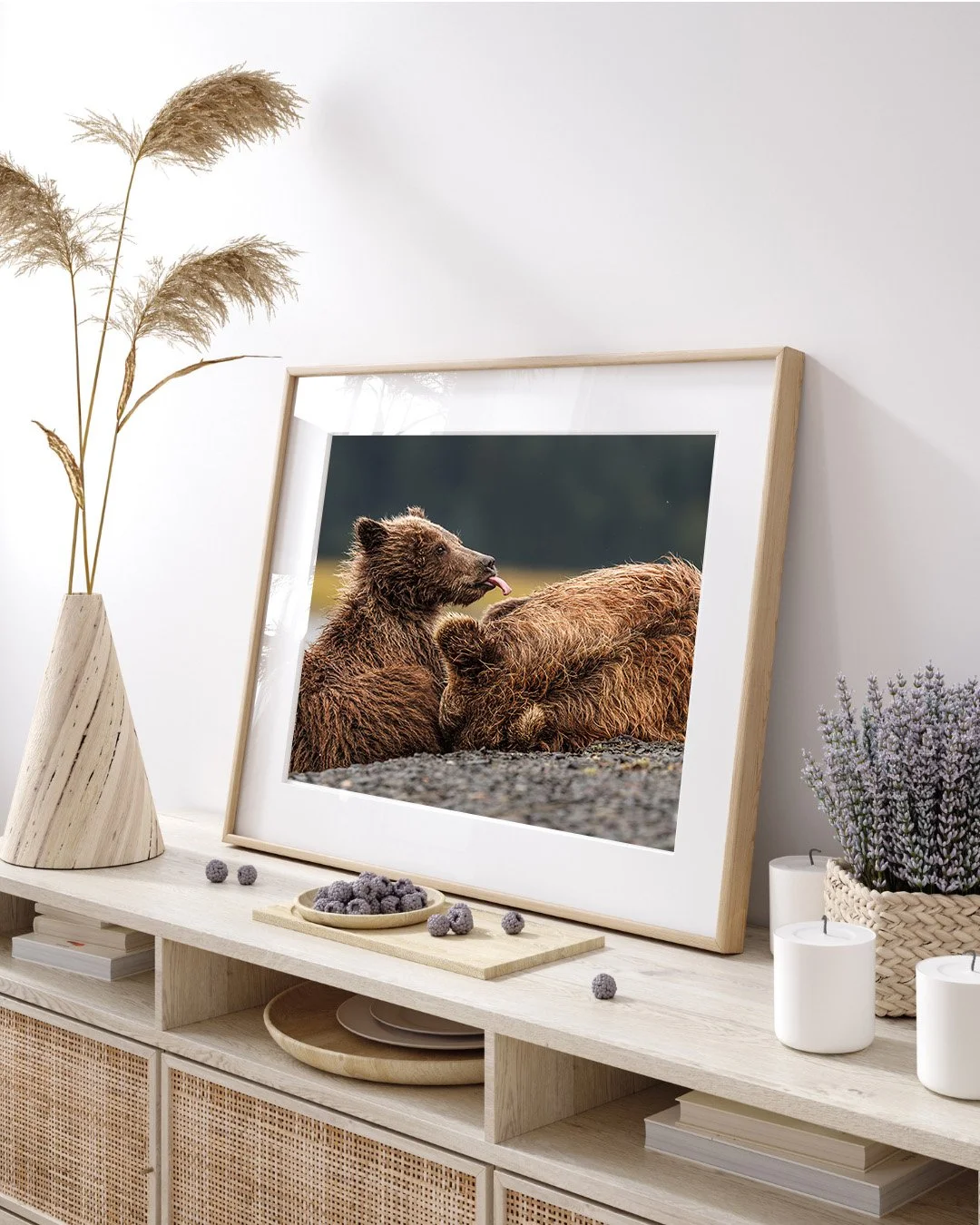 Framed photograph of two brown bears, one licking the other, on a wooden table decorated with white candles, a woven basket with purple flowers, and a light wooden vase with dried grass