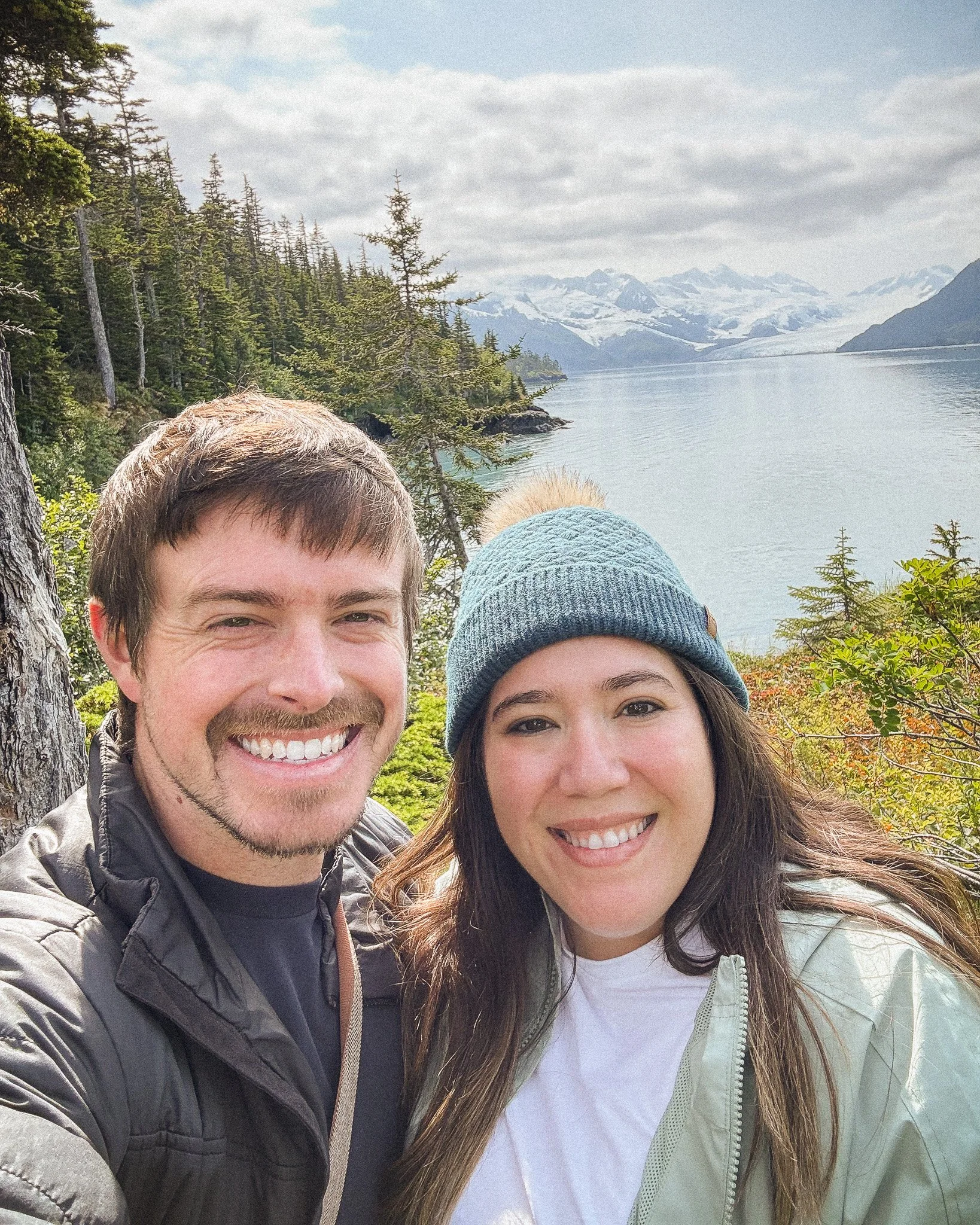 A smiling man and woman taking a selfie outdoors with a scenic mountain and lake background.