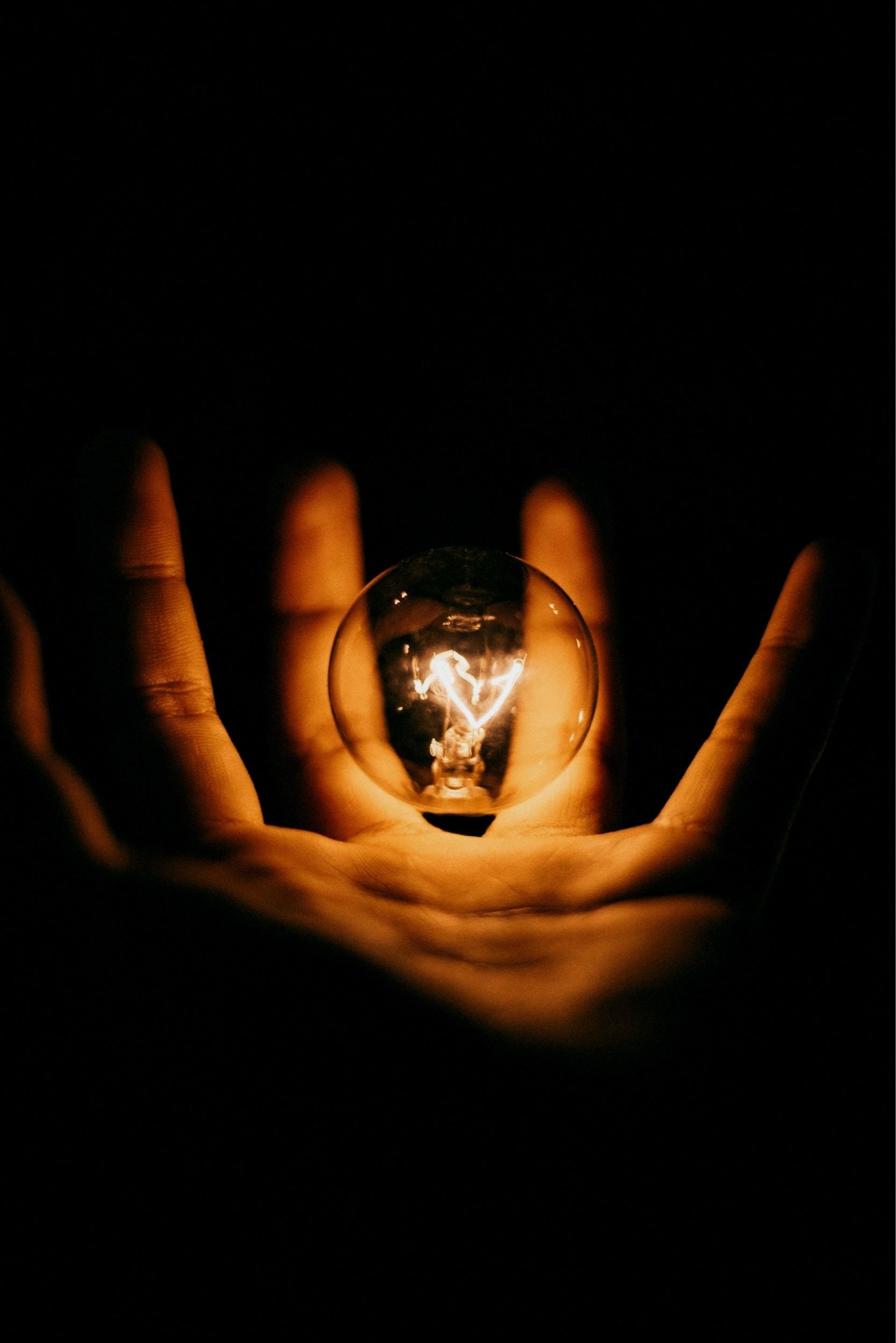 Hand holding a glowing light bulb with a heart-shaped filament inside, against a dark background.
