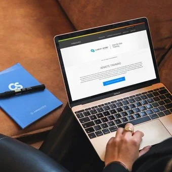 Person using a laptop with a screen displaying a request training webpage, a blue notebook, and a pen on a brown couch.