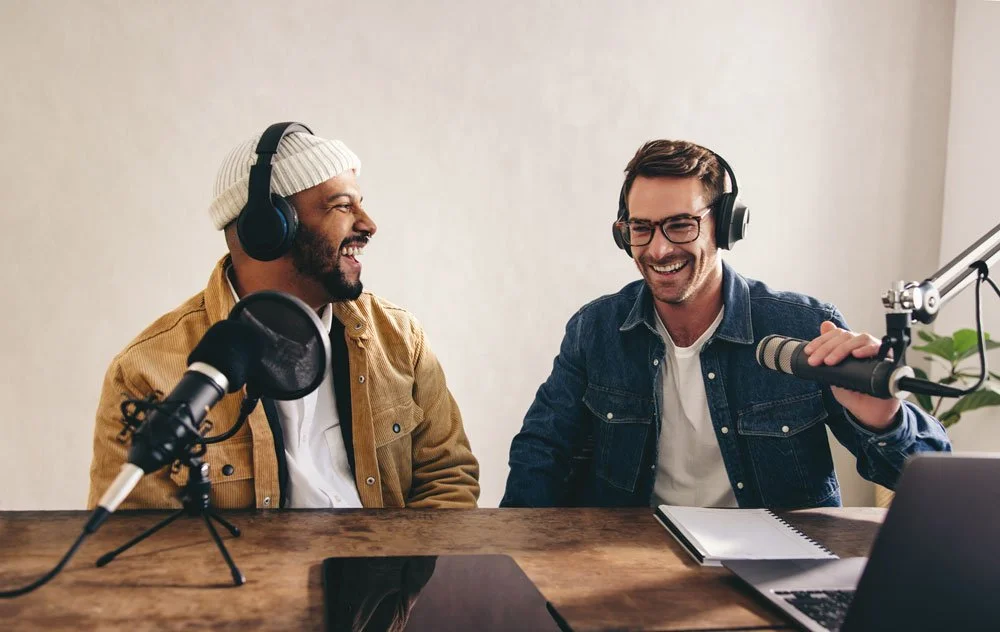 Two men wearing headphones are sitting at a table with microphones, smiling and talking, in a podcast recording setup.