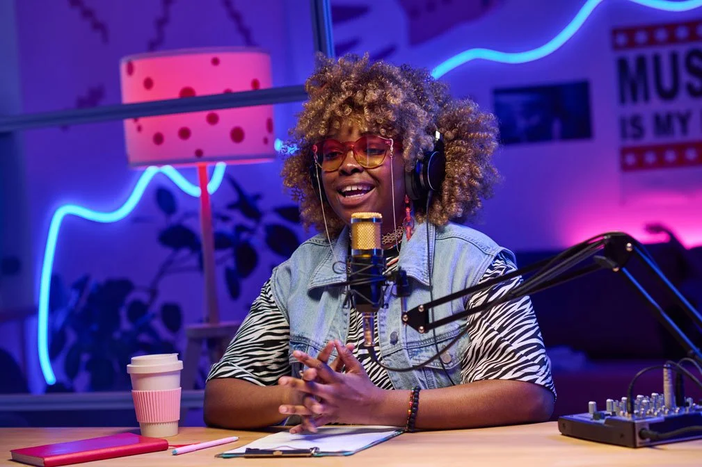 A woman with curly hair and glasses is speaking into a microphone on a podcast or radio show set, surrounded by colorful neon lights and music-themed decor.