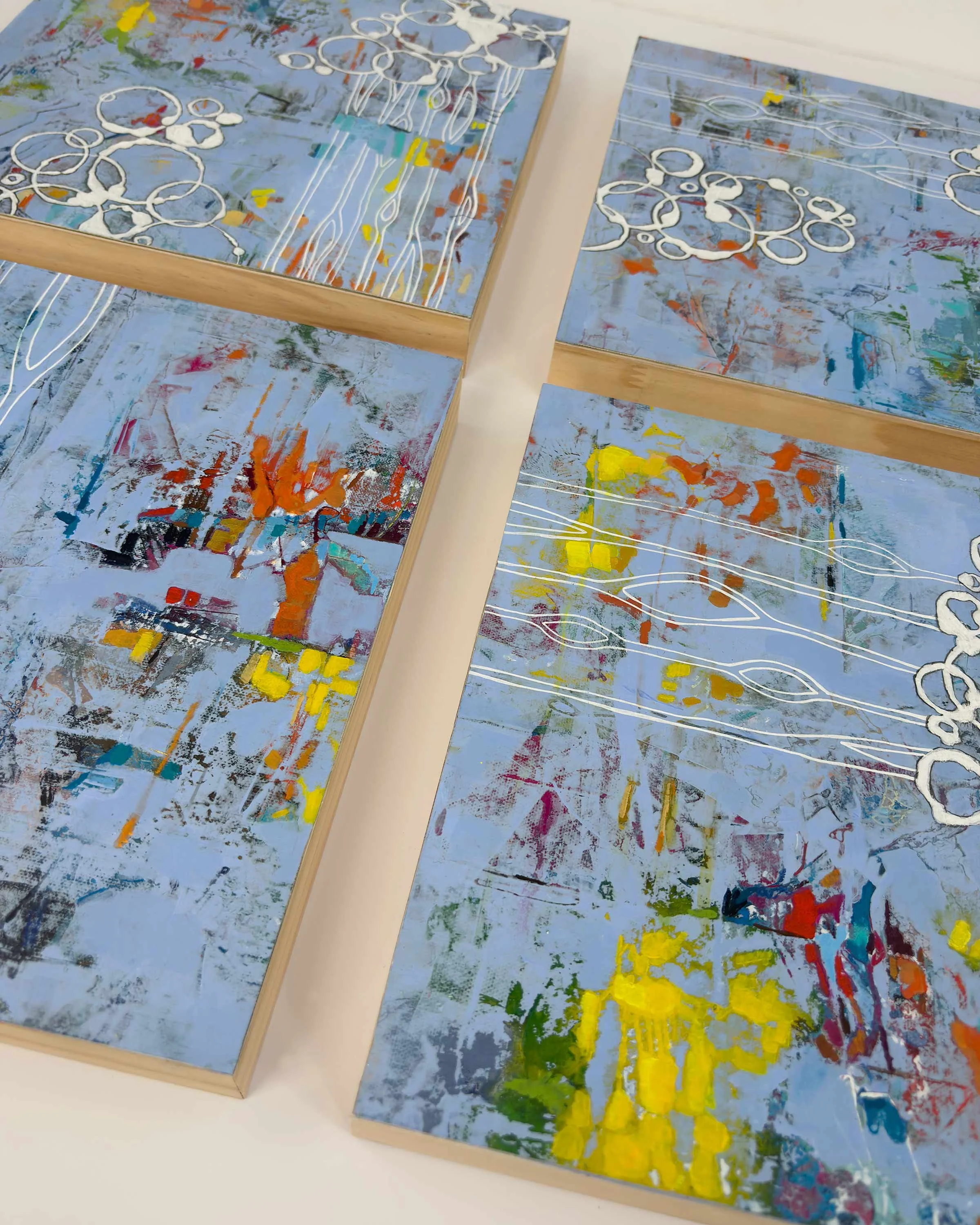 Overhead view of a four-piece abstract artwork on wood panels, with a calm blue base, expressive brushwork, and bright pops of color connected by white organic line patterns.
