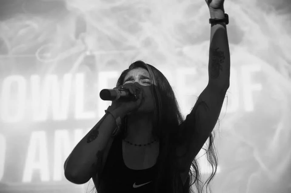 A woman with long dark hair singing into a microphone with her arm raised, in a black and white photo, likely at a concert or rally.